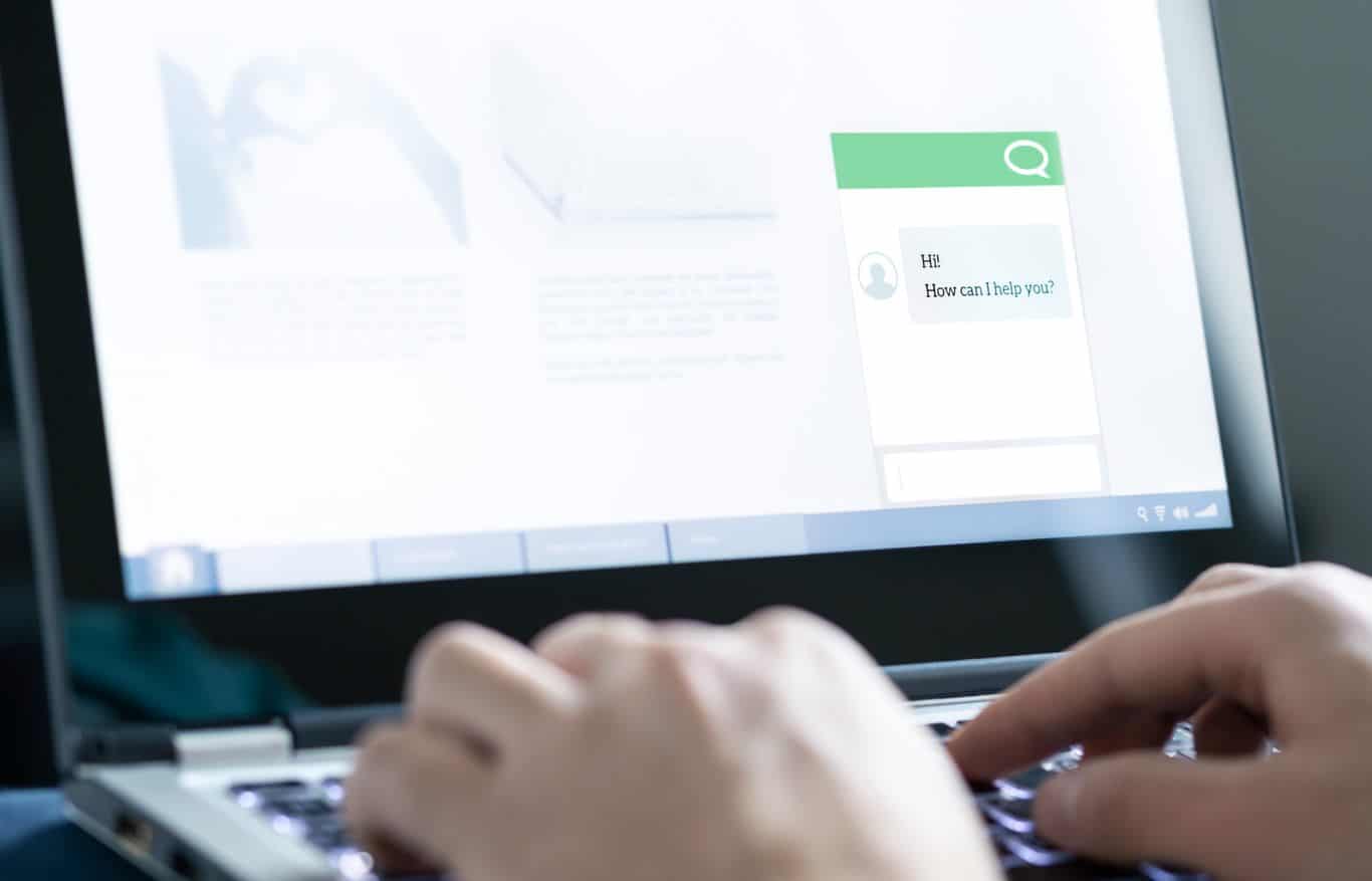 Image: Hands typing on a laptop with a screen that shows a chatbot asking 'how can we help?'